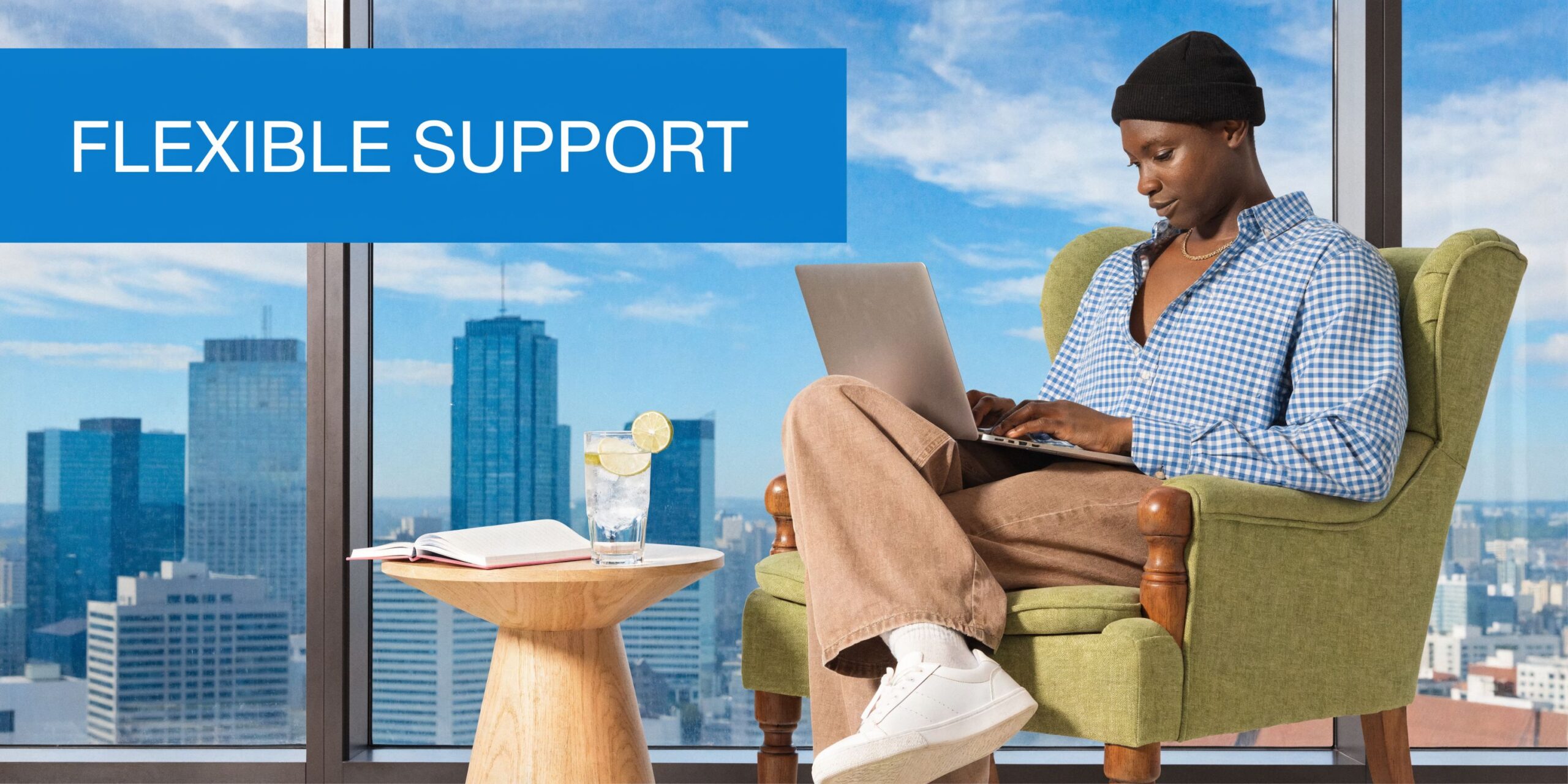 A person working on a laptop in a comfortable armchair with a city view and drink.