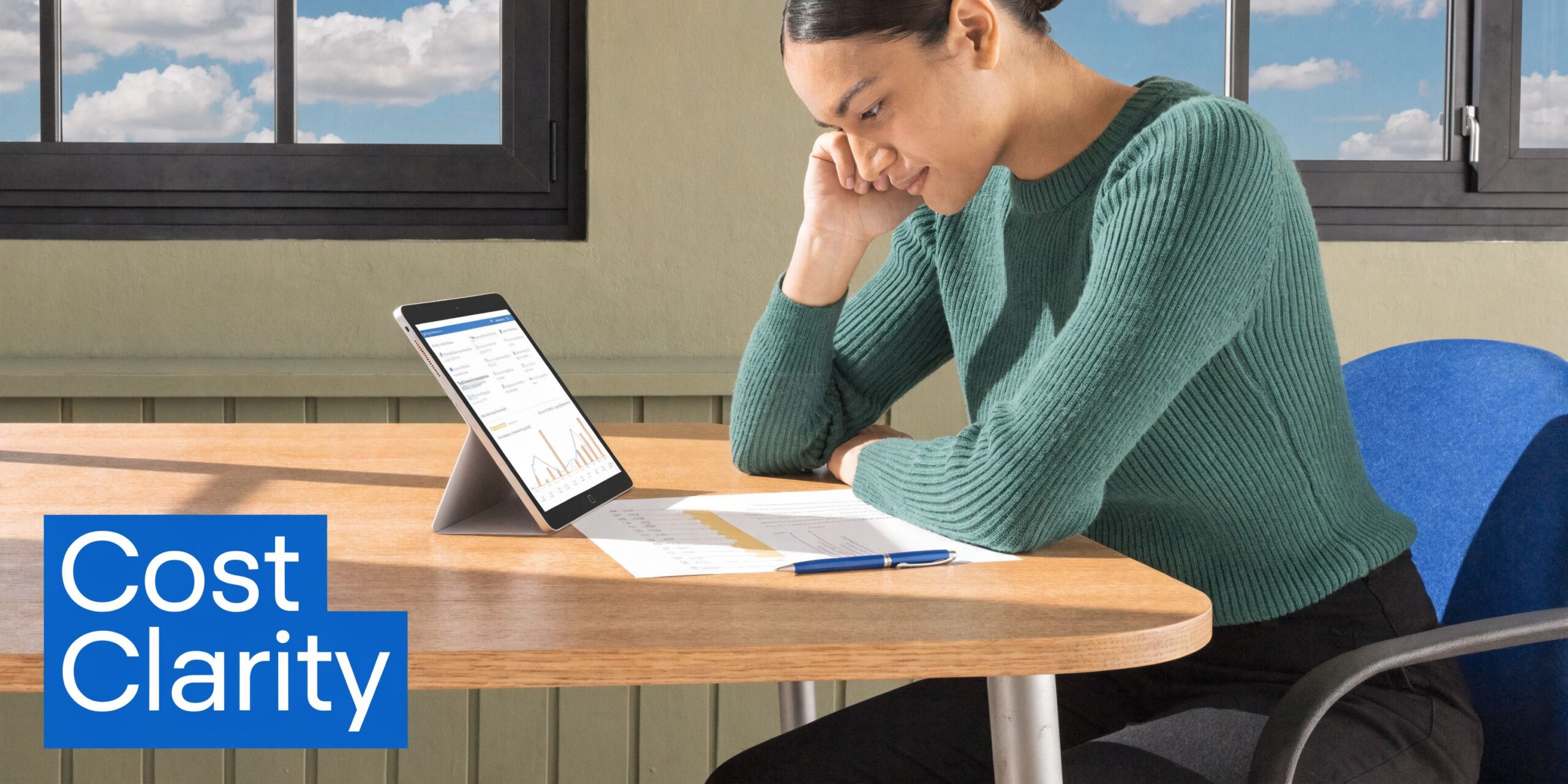 A woman reviewing financial data on a tablet and paper documents while seated at a wooden desk.