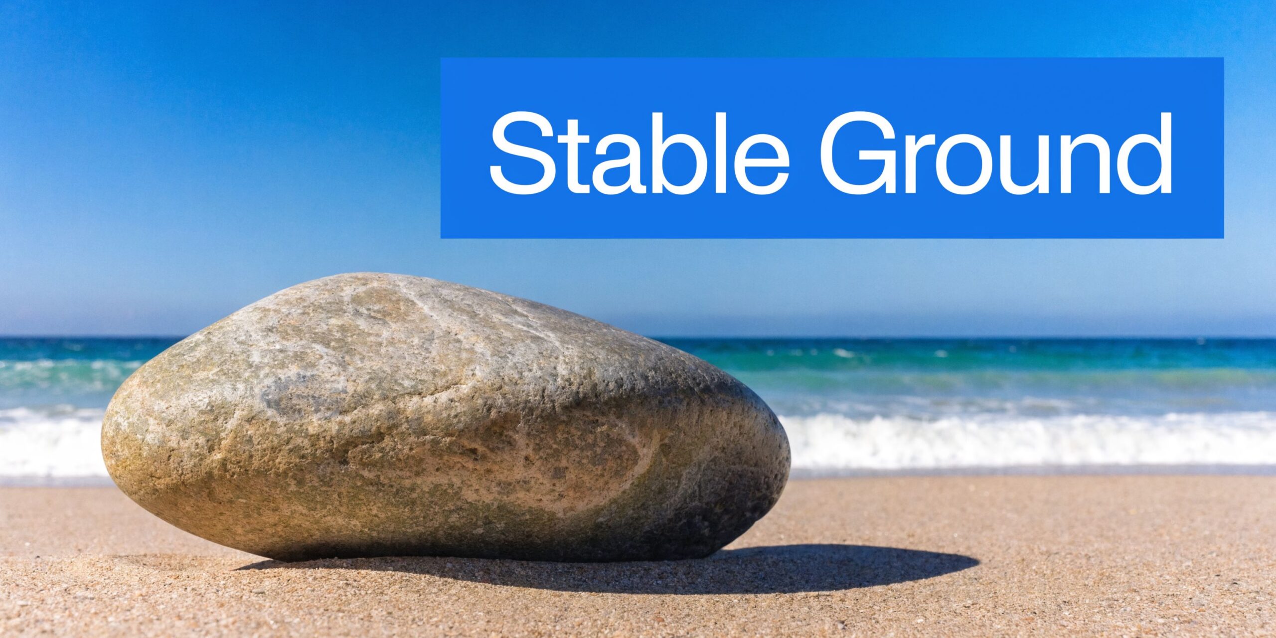 A large, smooth gray stone resting on a sandy beach with the ocean waves in the background.