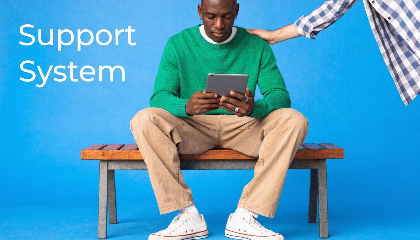 A man sitting on a bench looking at a tablet with someone placing a comforting hand on his shoulder.