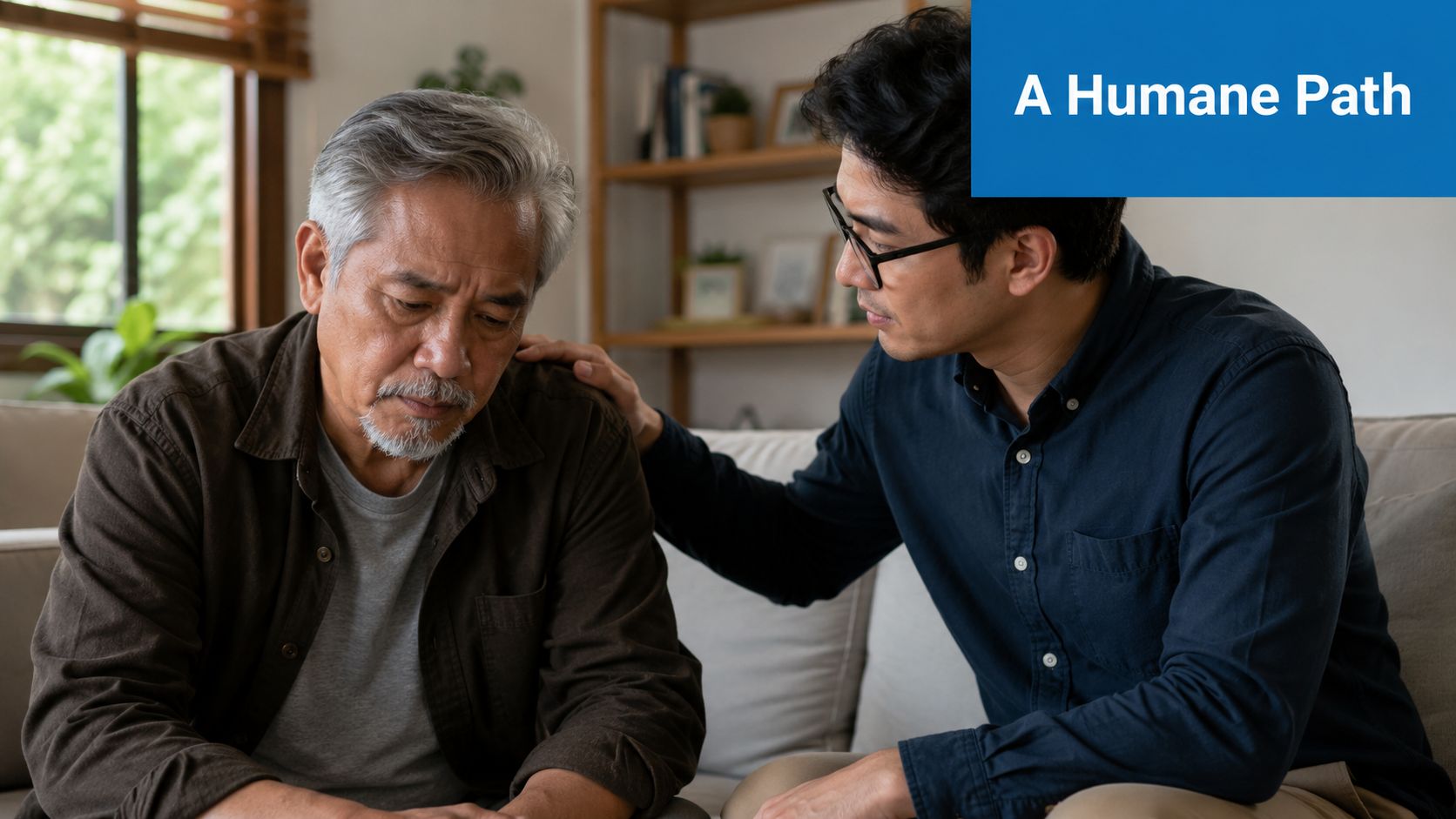 A younger Asian man offers comfort and support to an older man sitting on a couch.